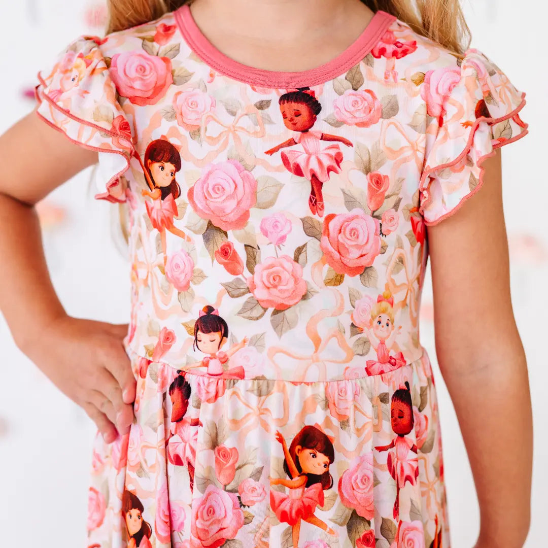 Close-up of a flutter-sleeve dress printed with pink roses, ribbons, and little ballerinas, with rose-pink neckline trim.